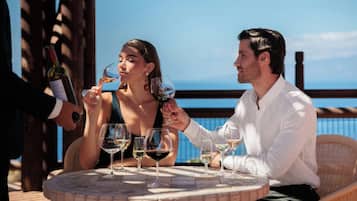 Two people seated at a round table with multiple wine glasses, overlooking a body of water.