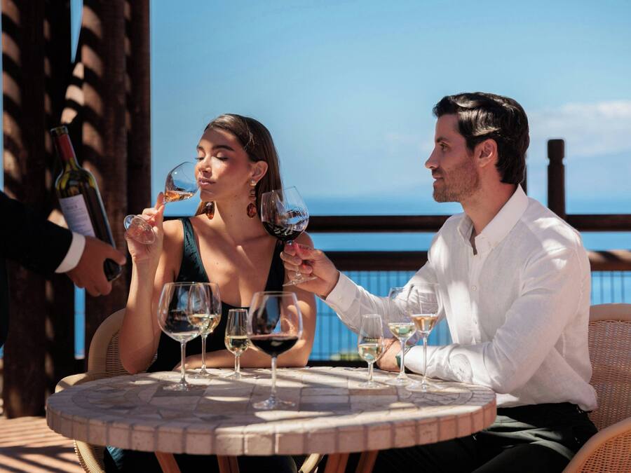 Two people seated at a round table with multiple wine glasses, overlooking a body of water.