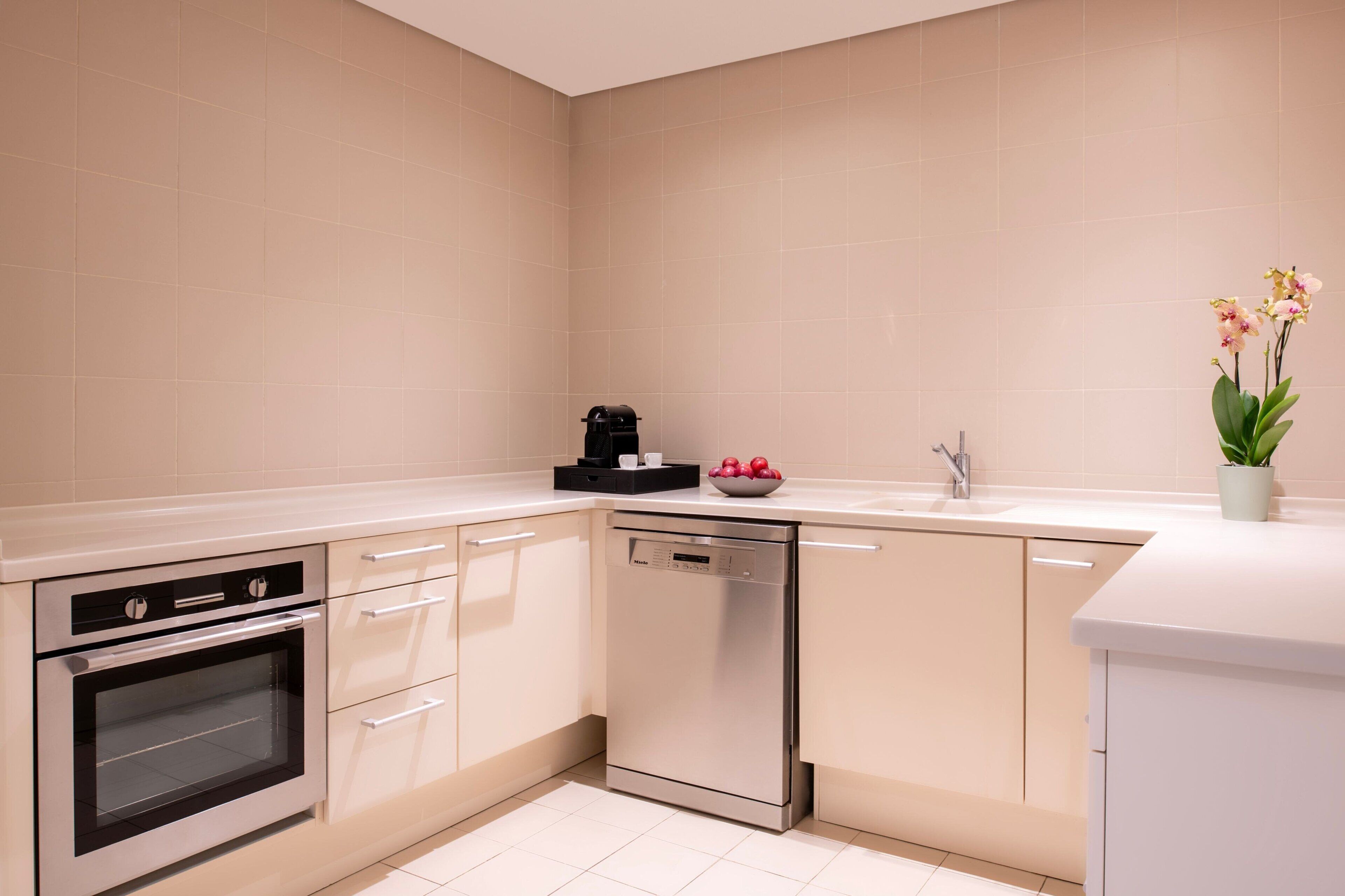 A modern kitchen with white cabinets, a built-in oven, a dishwasher, and a countertop with a coffee maker and a bowl of fruit.