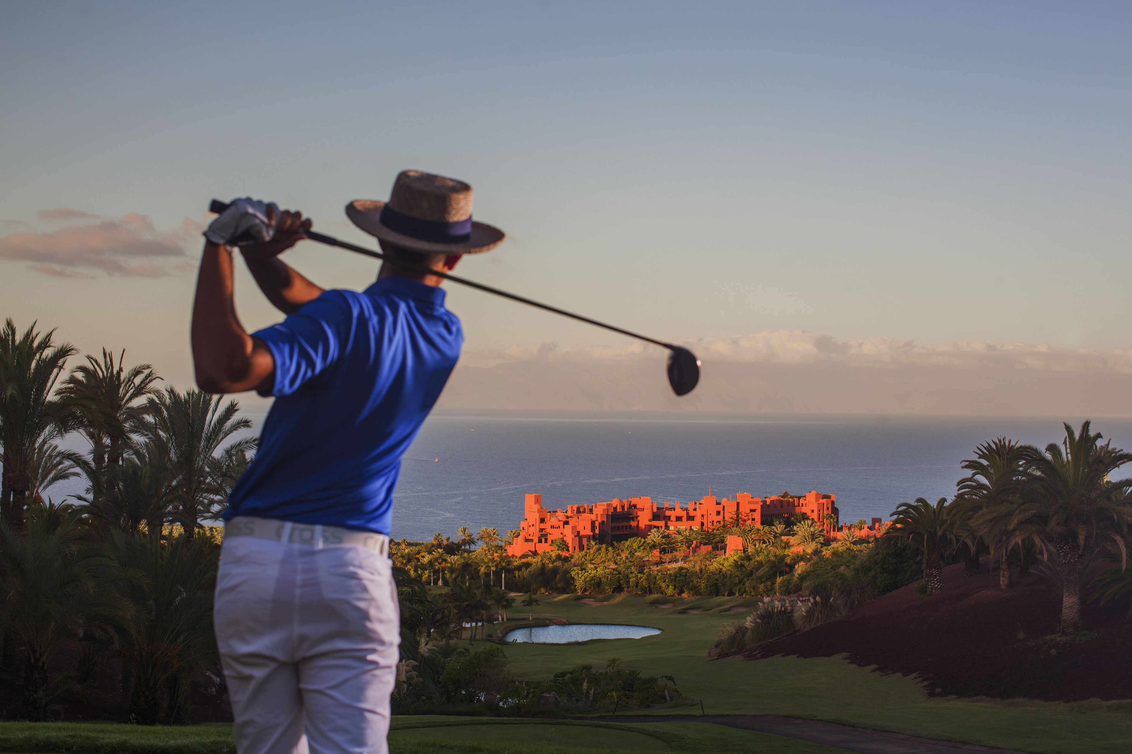 A golfer in a blue shirt and white pants preparing to swing, with a view of a coastal landscape and a large building in the background.