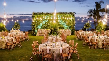 Una recepción de boda al aire libre con mesas redondas, sillas de mimbre y luces colgantes.