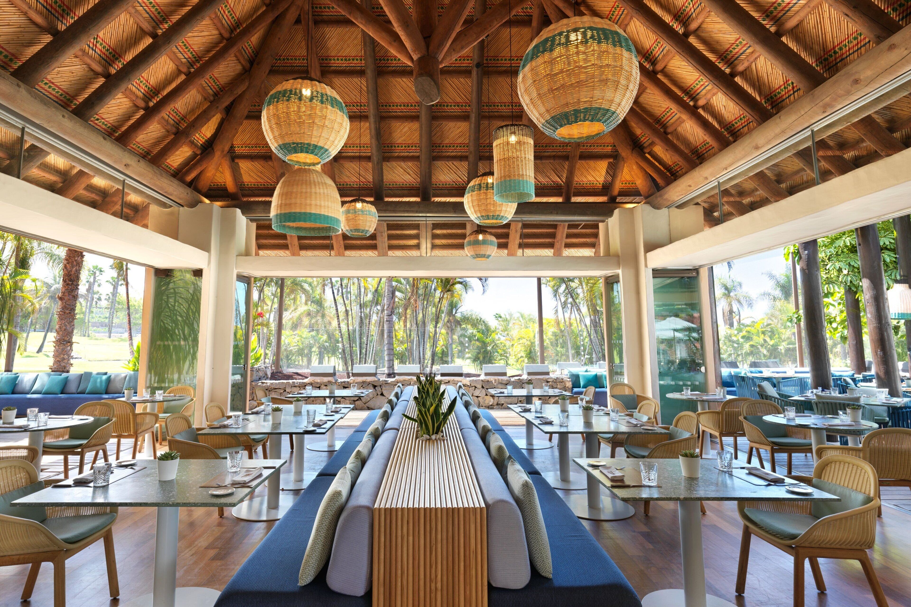 Un vaste espace extérieur aménagé pour les repas, avec des tables et des chaises en bois, un coin salon central et un toit de chaume.