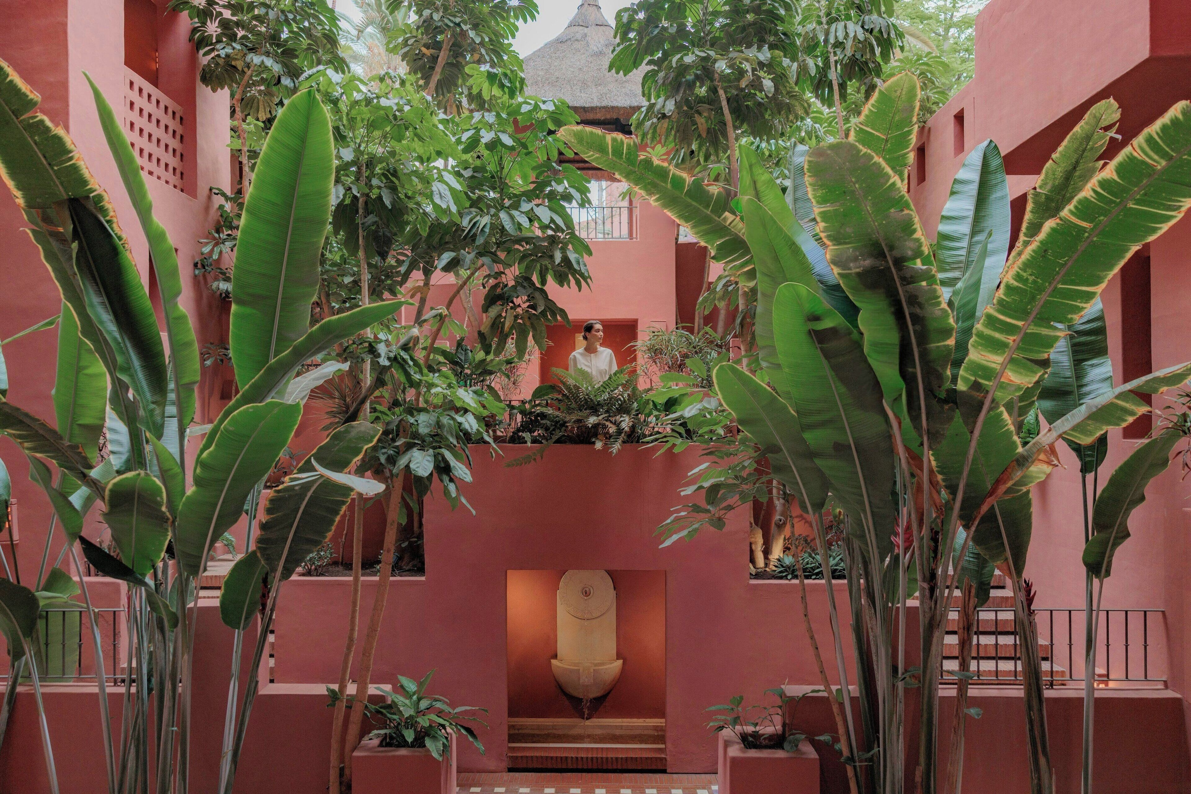 Un patio con una fuente central, rodeado de abundante vegetación y una fachada de edificio rosa.