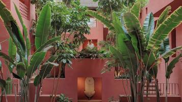 Un patio con una fuente central, rodeado de abundante vegetación y una fachada de edificio rosa.