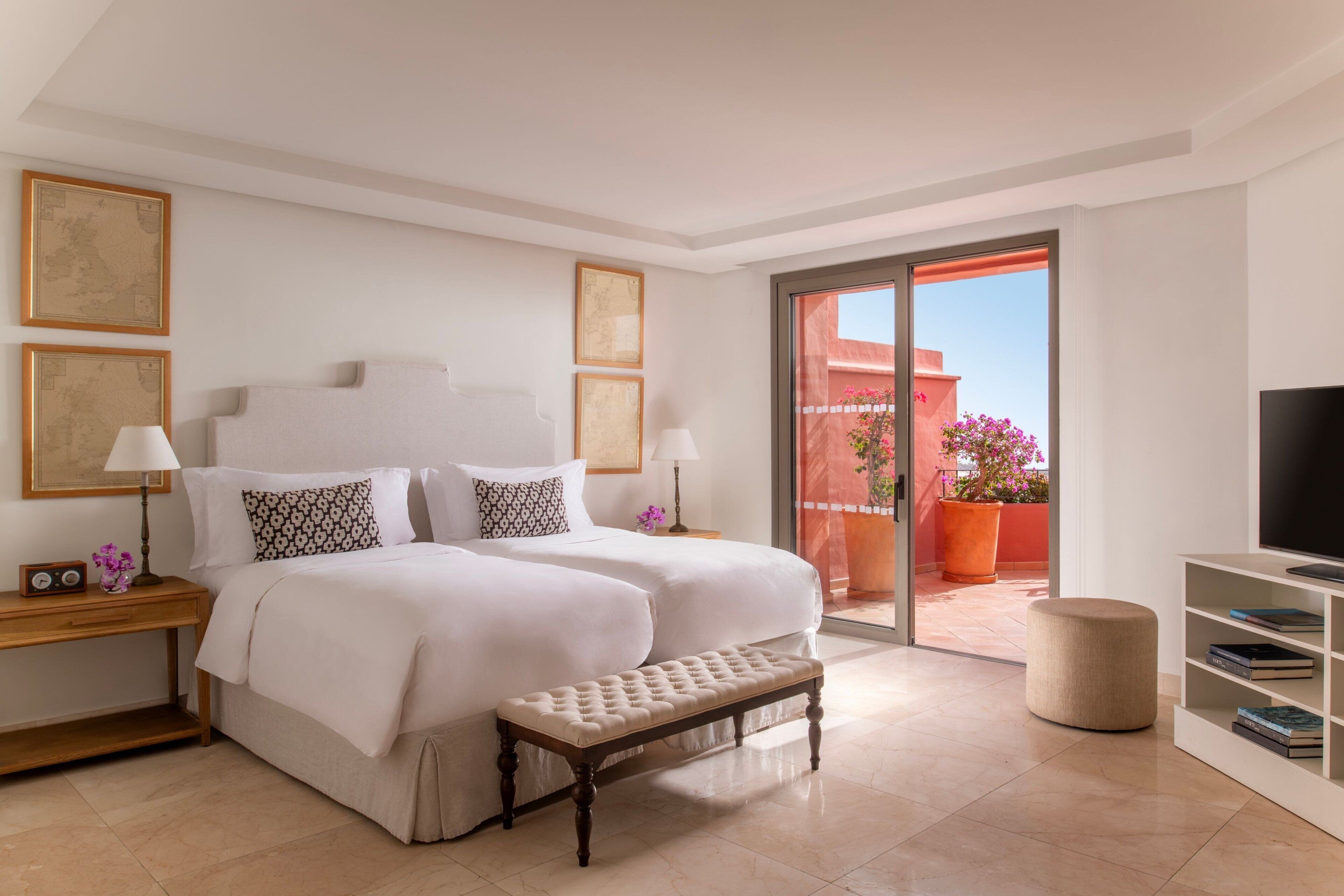 A hotel room with two beds, a bench, a TV, and a balcony with a potted plant.
