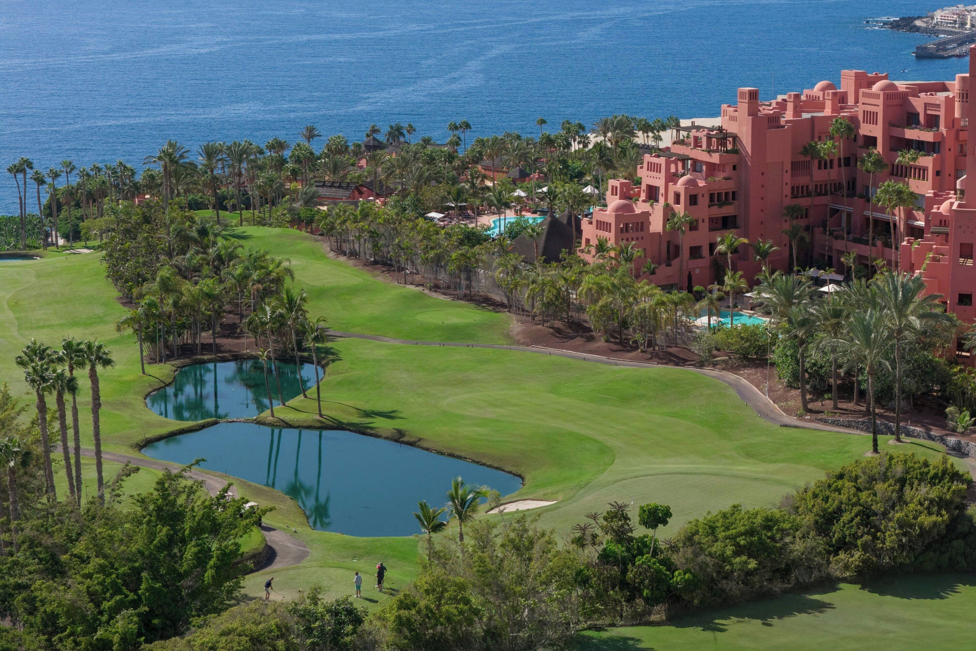 A golf course with a pond, palm trees, and a large building with a pool.