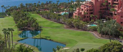 A golf course with a pond, palm trees, and a large building with a pool.