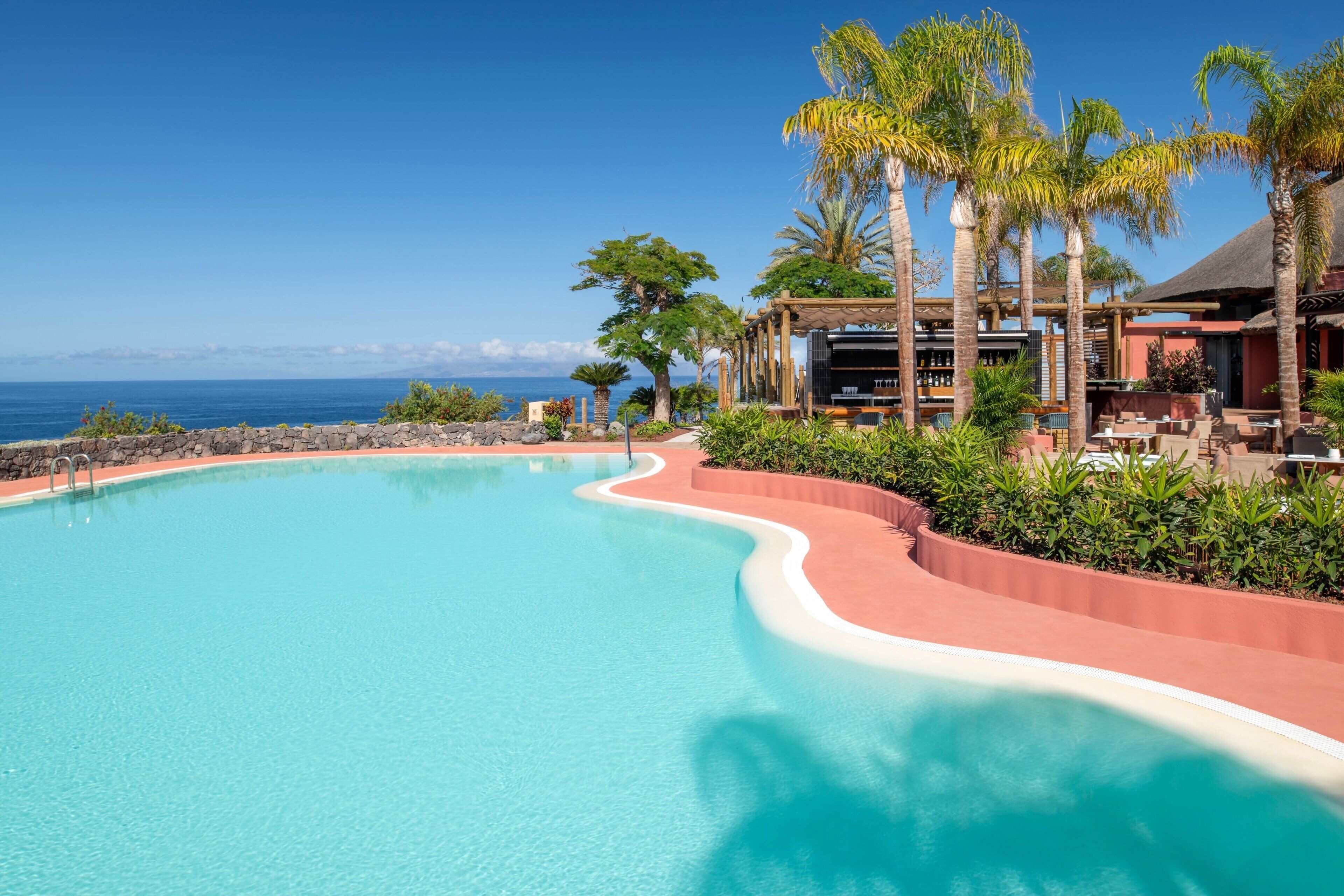 Piscina de resort con borde curvo, rodeada de palmeras y vista al océano.