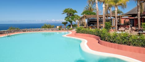 A resort pool with a curved edge, surrounded by palm trees and a view of the ocean.