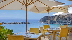 An outdoor dining area with wicker chairs and tables, overlooking a coastal view with a rocky cliff and the sea.