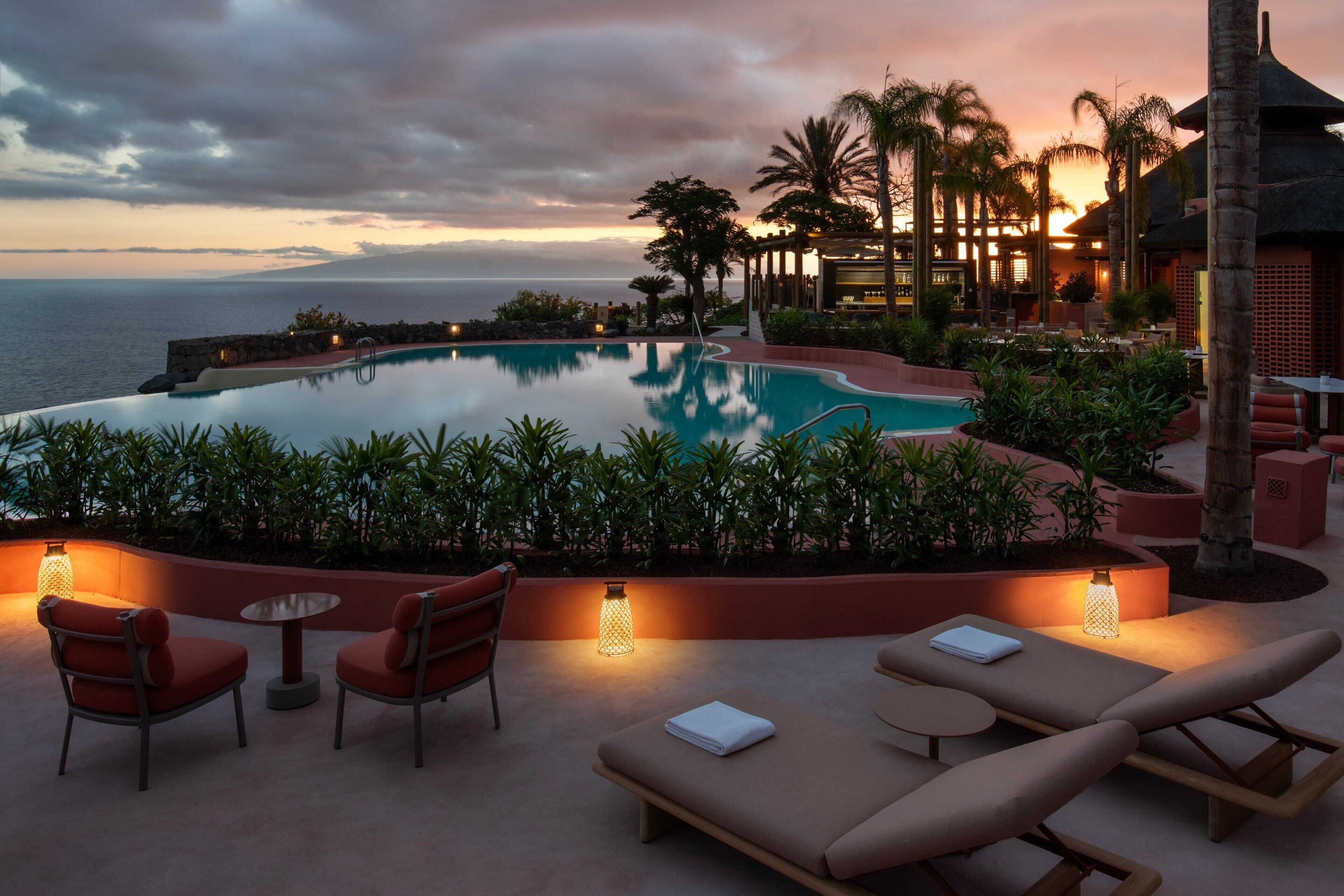 An infinity pool with a view of the ocean at sunset, surrounded by palm trees and lounge chairs.