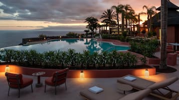 An infinity pool with a view of the ocean at sunset, surrounded by palm trees and lounge chairs.