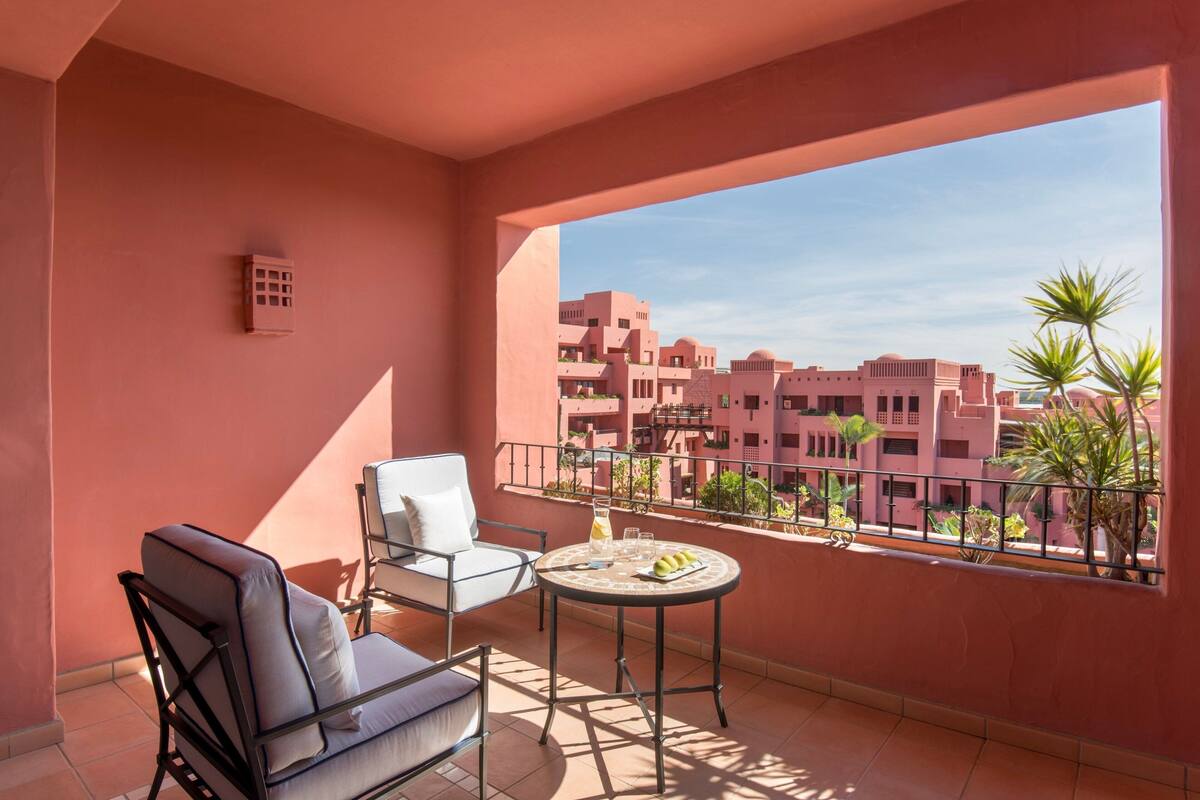Un balcón con vistas a edificios rosados, una mesa con una bebida y dos sillas.