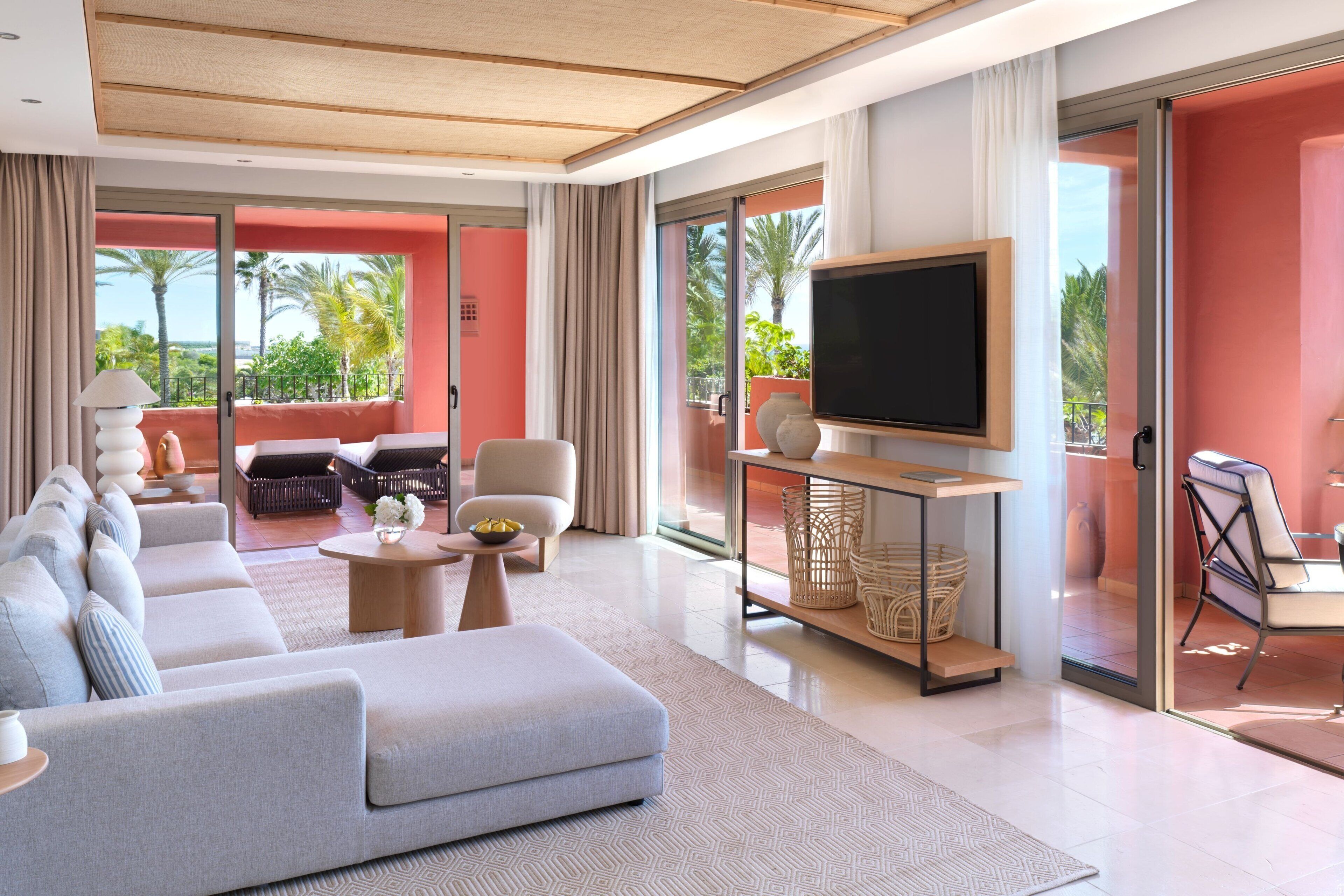 A modern living room with a large grey sofa, a flat-screen TV, and a balcony with a view of palm trees.