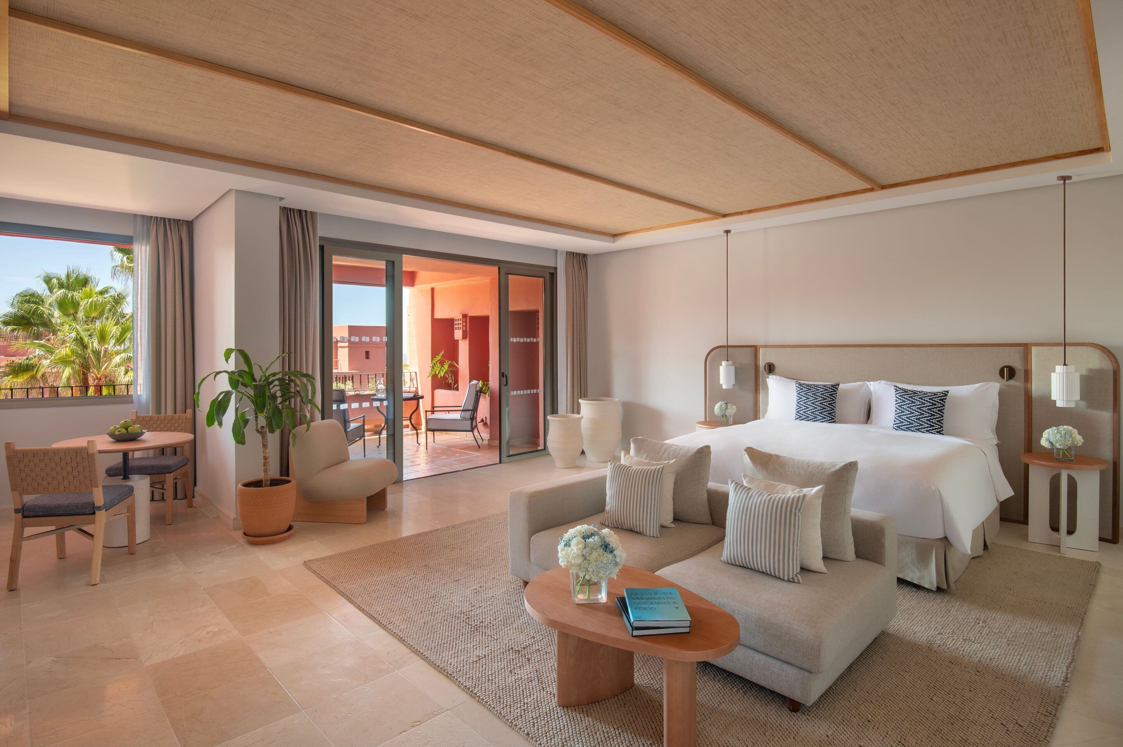 A modern hotel room with a large bed, a sofa, a dining area, and a balcony with a view of palm trees.