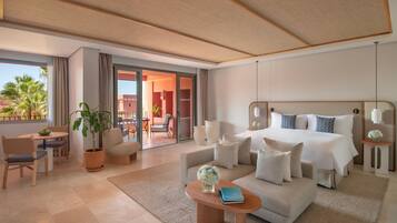 A modern hotel room with a large bed, a sofa, a dining area, and a balcony with a view of palm trees.