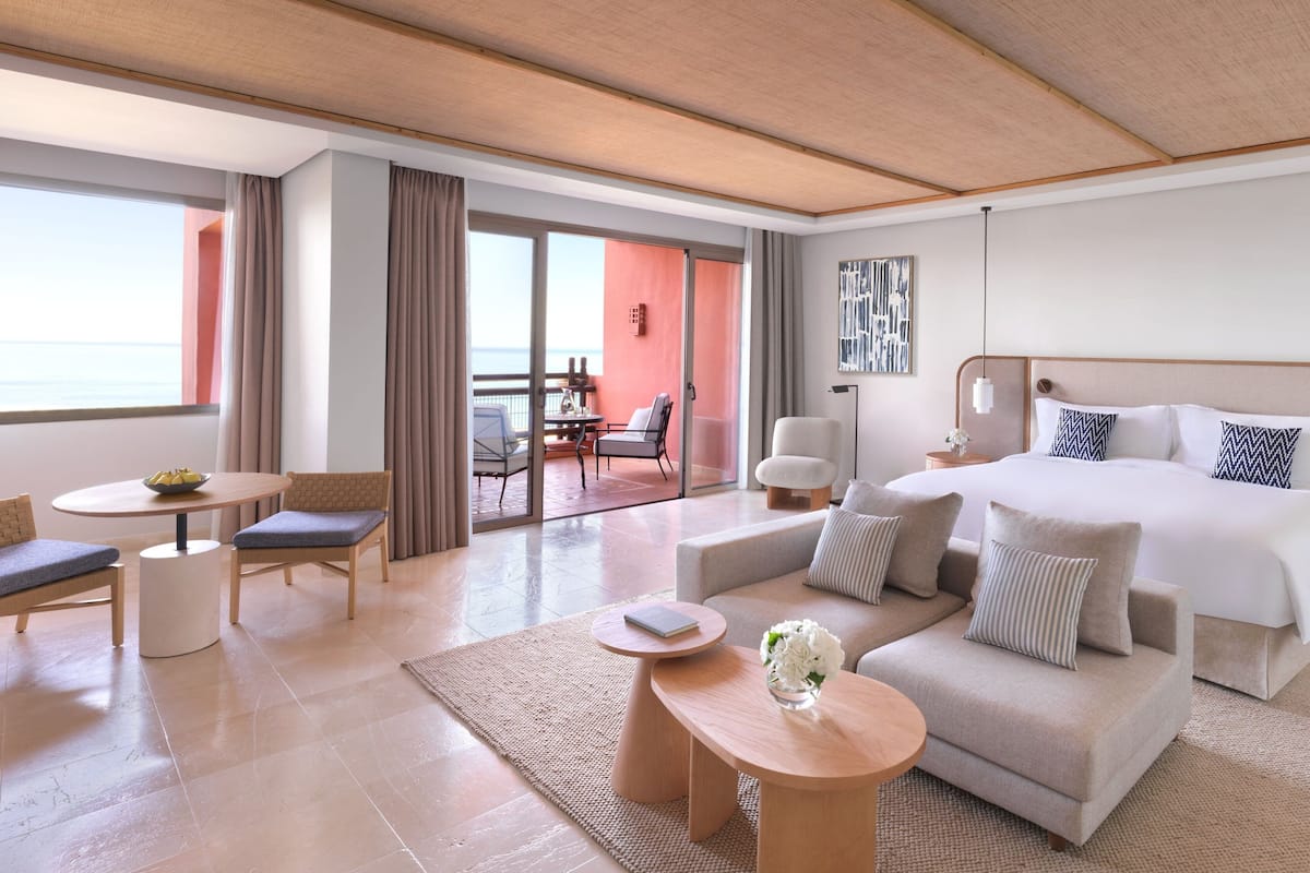 A modern hotel room with a large bed, a sofa, a dining table, and a balcony with a view of the sea.