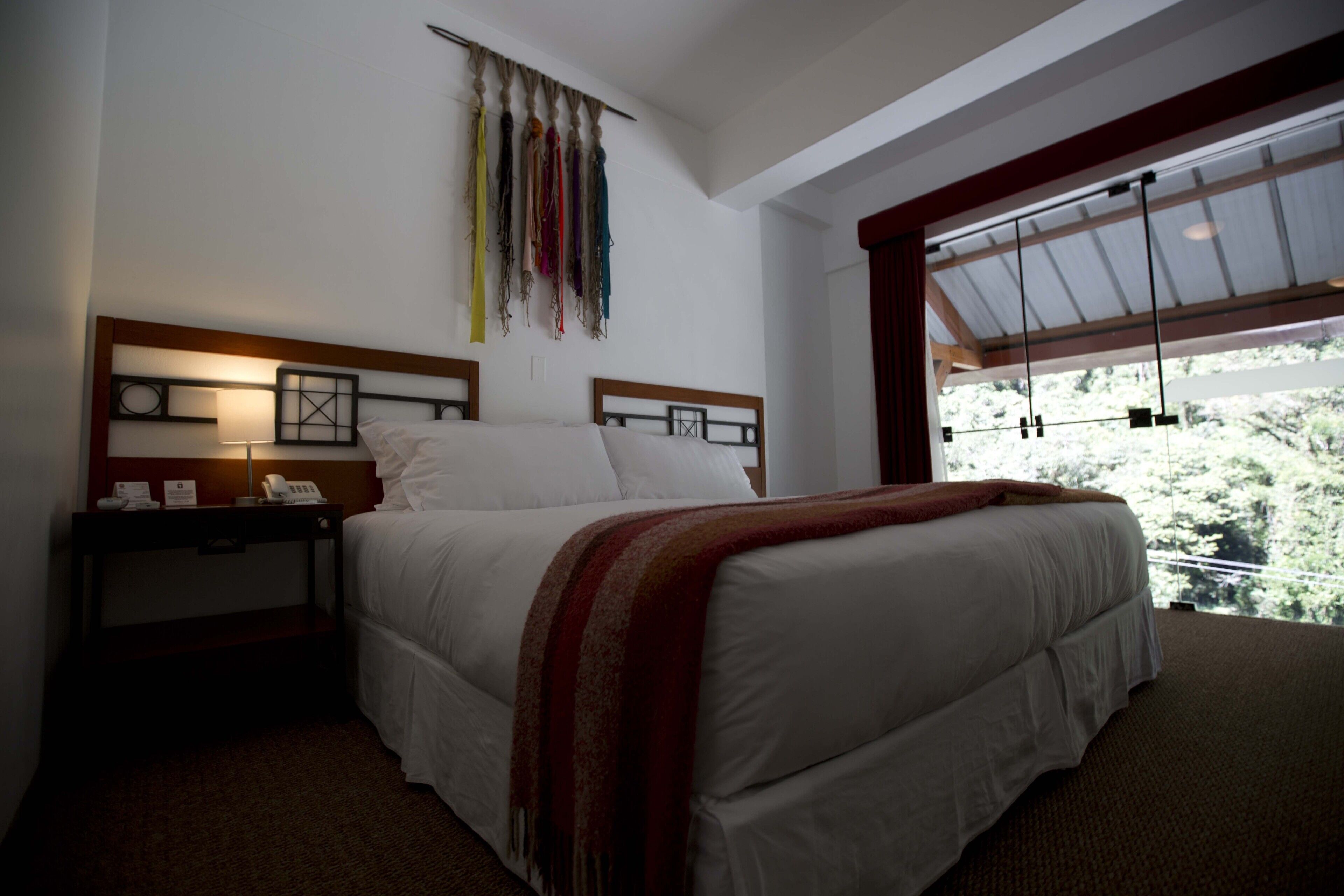 A hotel room with a large bed, a nightstand with a lamp, and a view of trees through a large window.