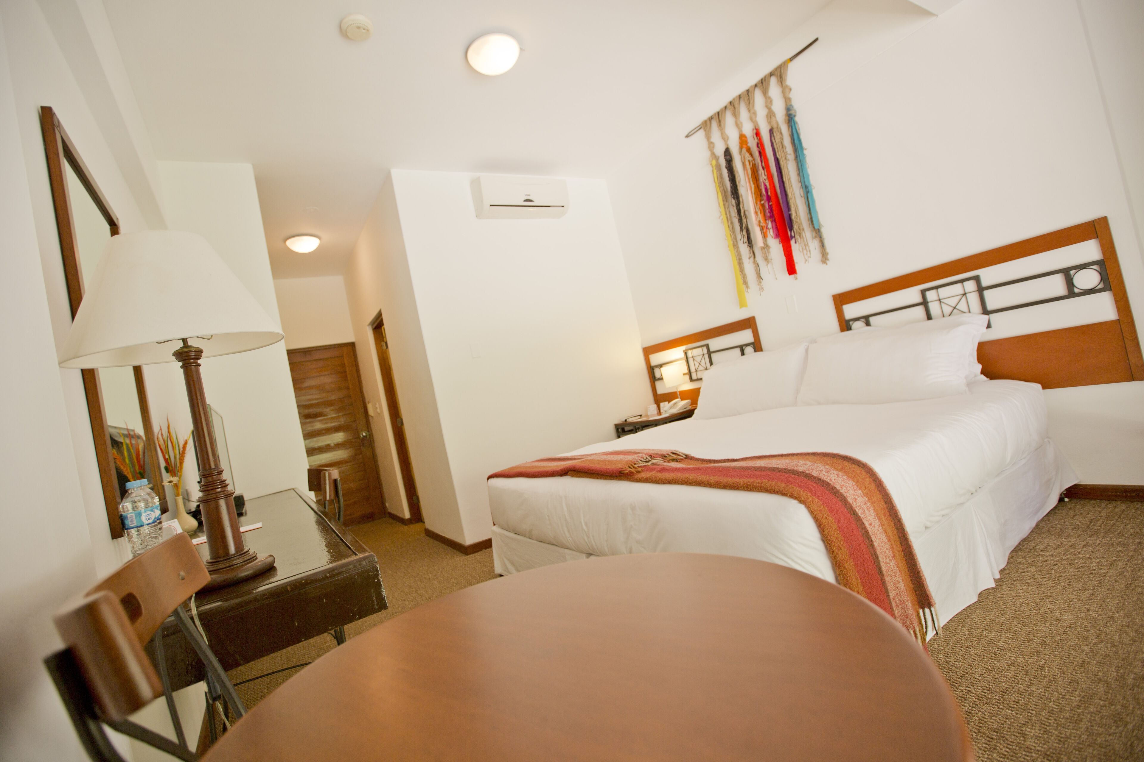 A hotel room with a bed, a desk with a lamp, a chair, and a wall-mounted air conditioner.