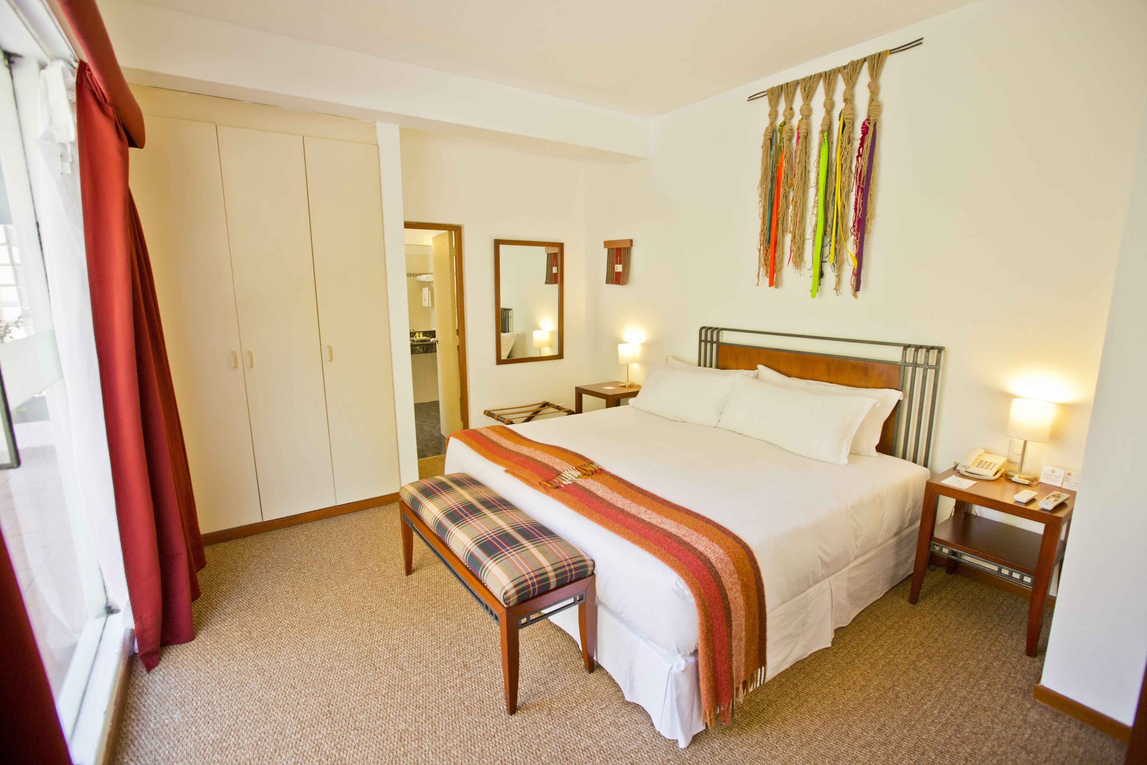 A hotel room with a large bed, a bench, a bedside table with a lamp, a mirror, and a wardrobe.