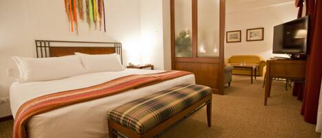 A hotel room with a large bed, a bench, a television, and a desk.