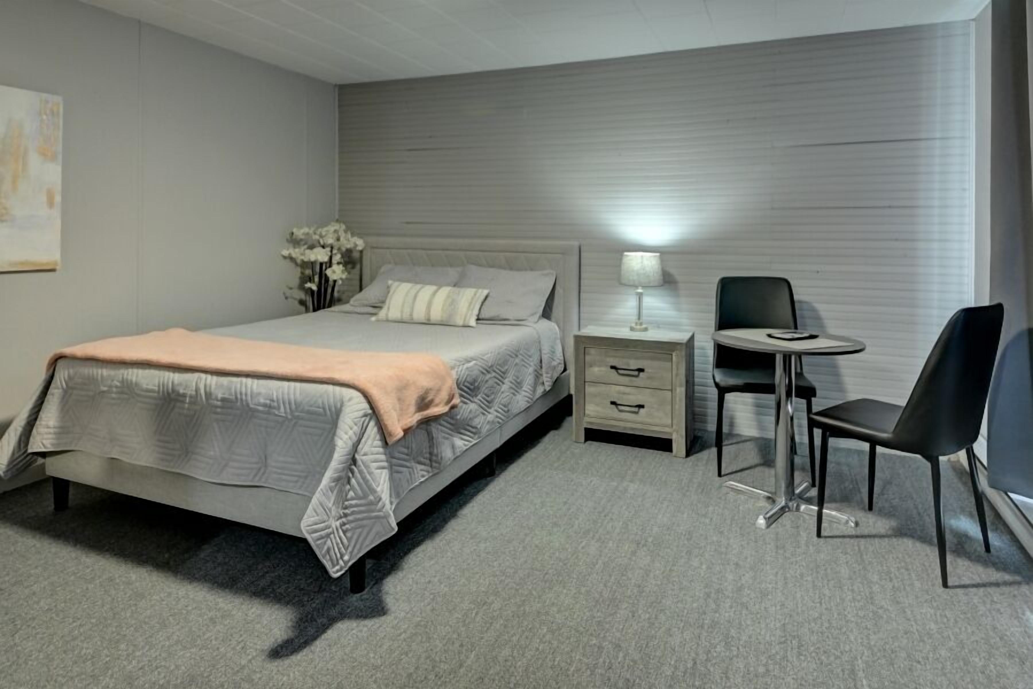 A bedroom with a bed, bedside table, lamp, chair, and a small table.
