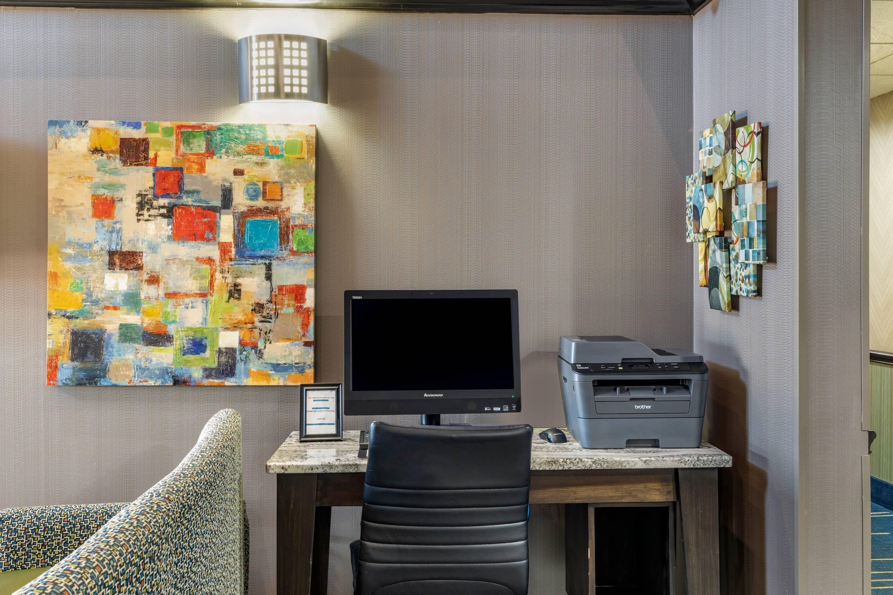 Une chambre d’hôtel équipée d’un bureau, d’un ordinateur, d’une imprimante, d’une chaise, d’un tableau abstrait aux couleurs vives et d’une œuvre d’art fixée au mur.