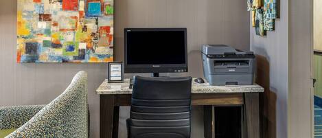 A hotel room with a desk, a computer, a printer, a chair, a colorful abstract painting, and a wall-mounted art piece.