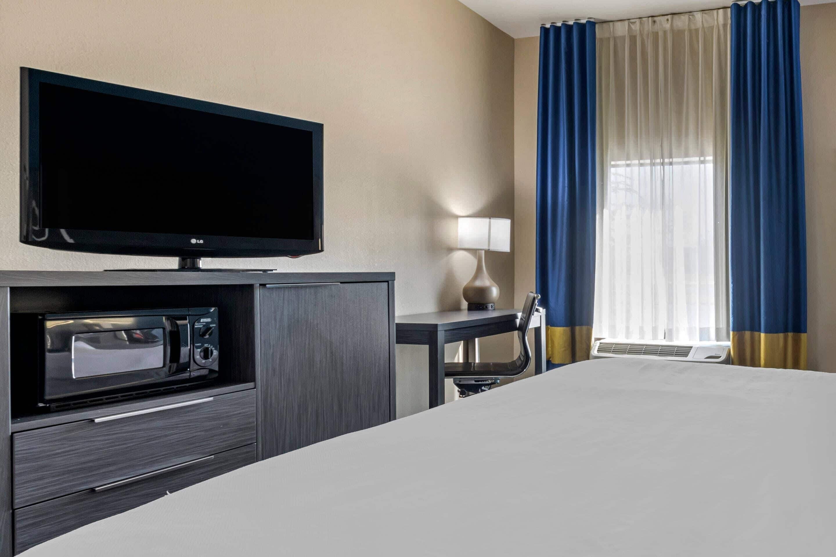 A hotel room with a bed, a flat-screen TV, a microwave, a desk with a lamp, and a window with blue curtains.