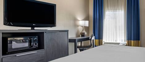 A hotel room with a bed, a flat-screen TV, a microwave, a desk with a lamp, and a window with blue curtains.