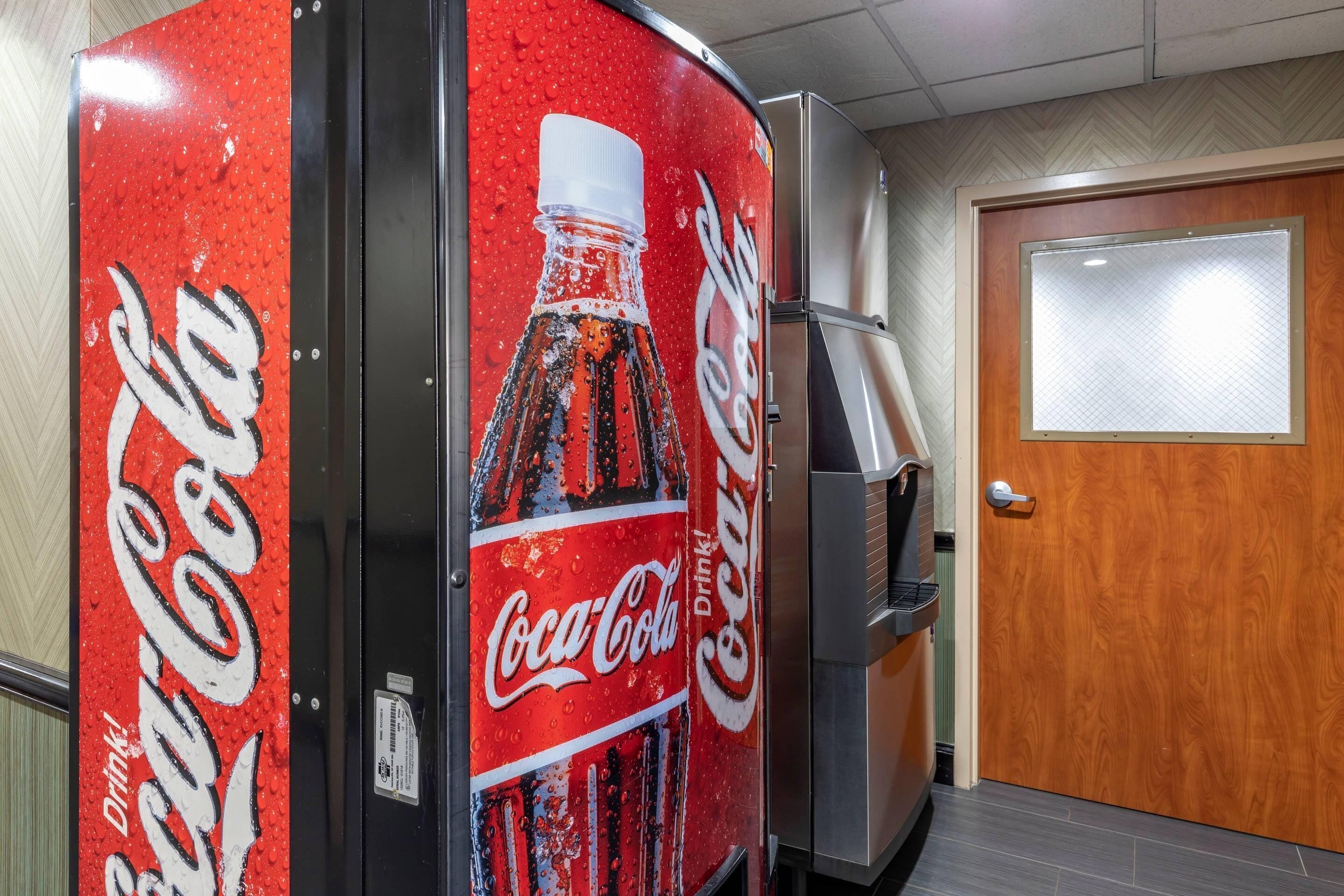 Un distributeur automatique de Coca-Cola se trouve dans une pièce dotée d’une porte en bois et d’un sol carrelé.
