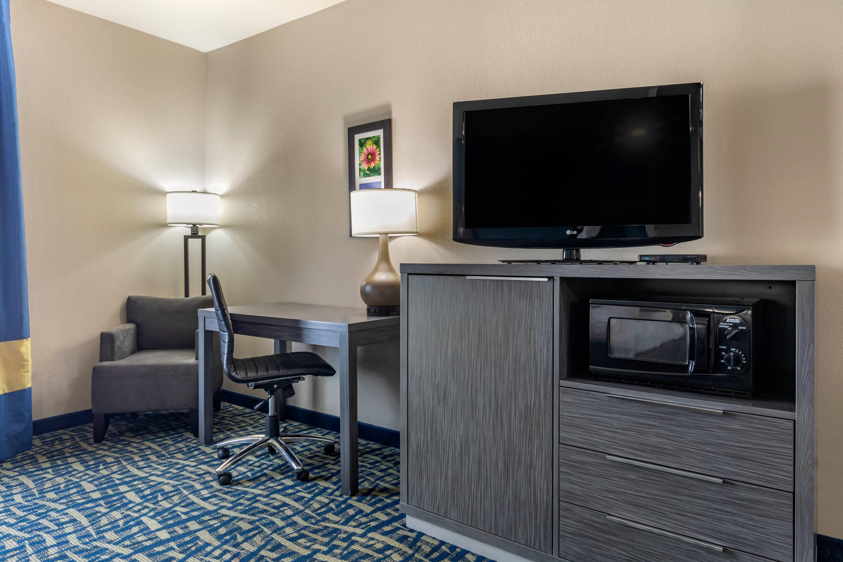 A hotel room with a flat-screen TV, a microwave, a desk with a chair, and a lamp.