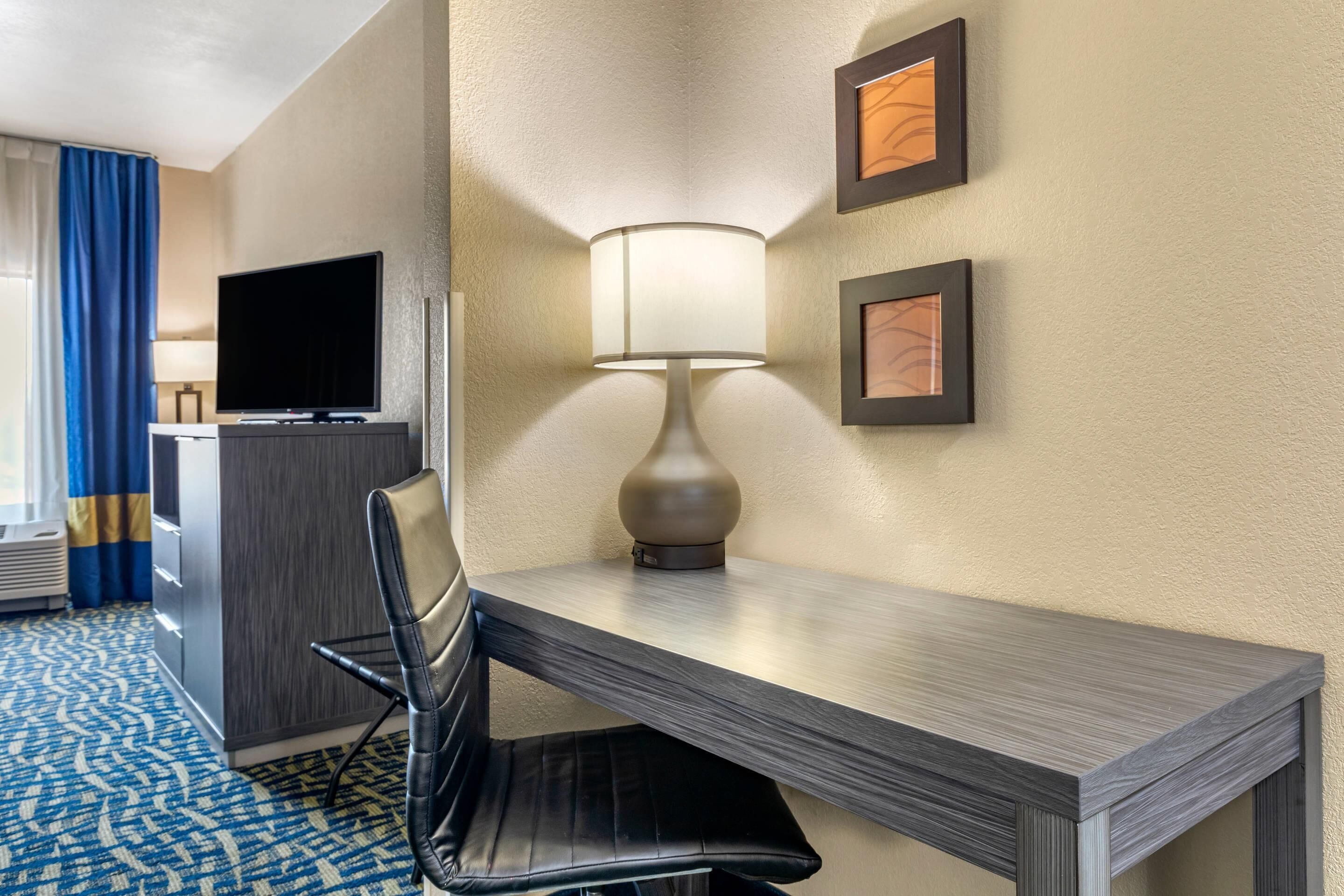 A hotel room with a desk, chair, TV, lamp, and artwork on the wall.