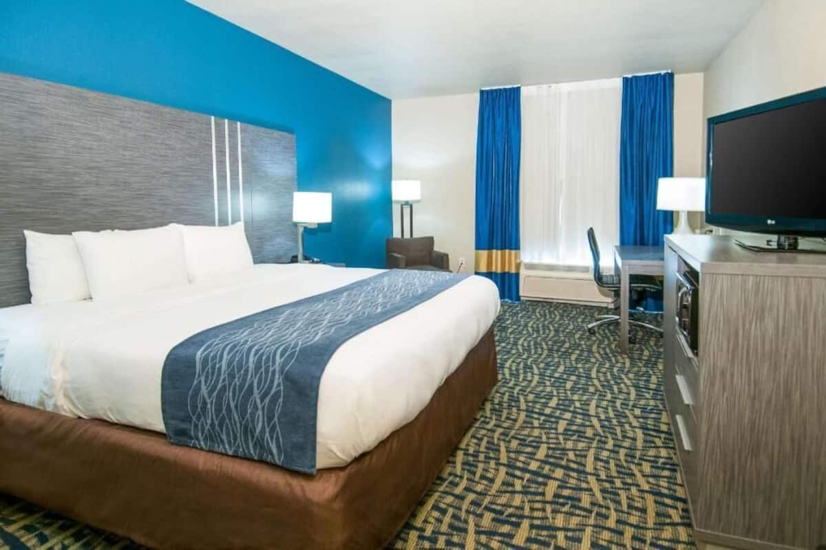 Ein Hotelzimmer mit einem großen Bett, einem Schreibtisch mit Computer, einem Fernseher und einem Fenster mit Vorhängen.