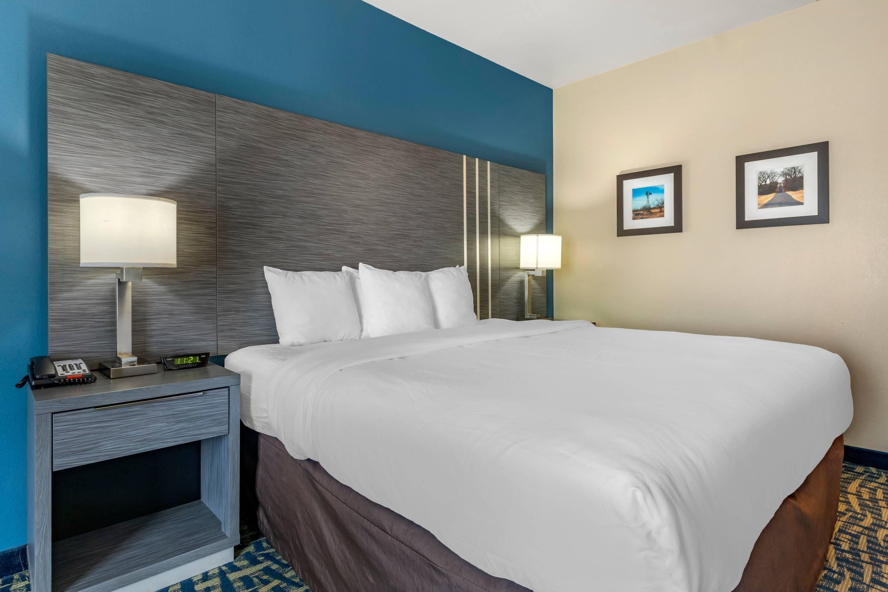 A hotel room with a large bed, two bedside lamps, a nightstand with a phone and a digital clock, and two framed pictures on the wall.