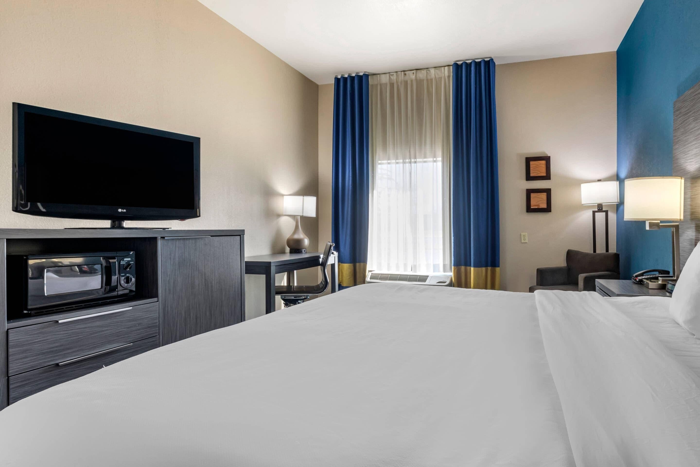 A hotel room with a bed, a flat-screen TV, a microwave, a desk with a lamp, and a window with curtains.
