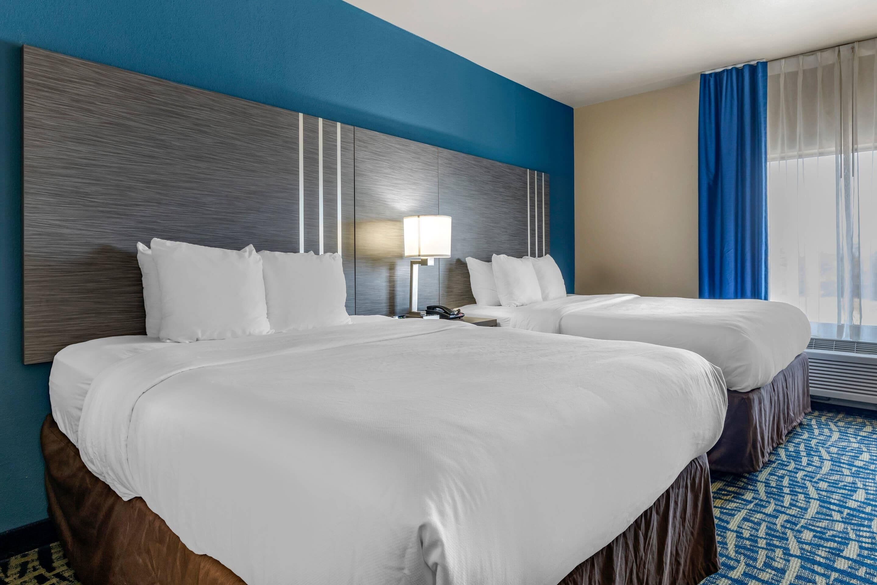Una habitación de hotel con dos camas, un cabecero grande y adosado a la pared, una mesita de noche con lámpara y una ventana con cortinas azules.