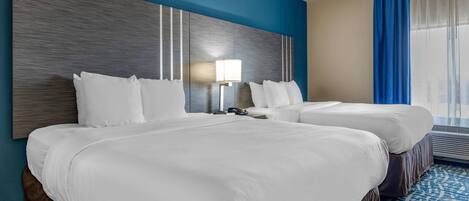 A hotel room with two beds, a large wall-mounted headboard, a bedside table with a lamp, and a window with blue curtains.