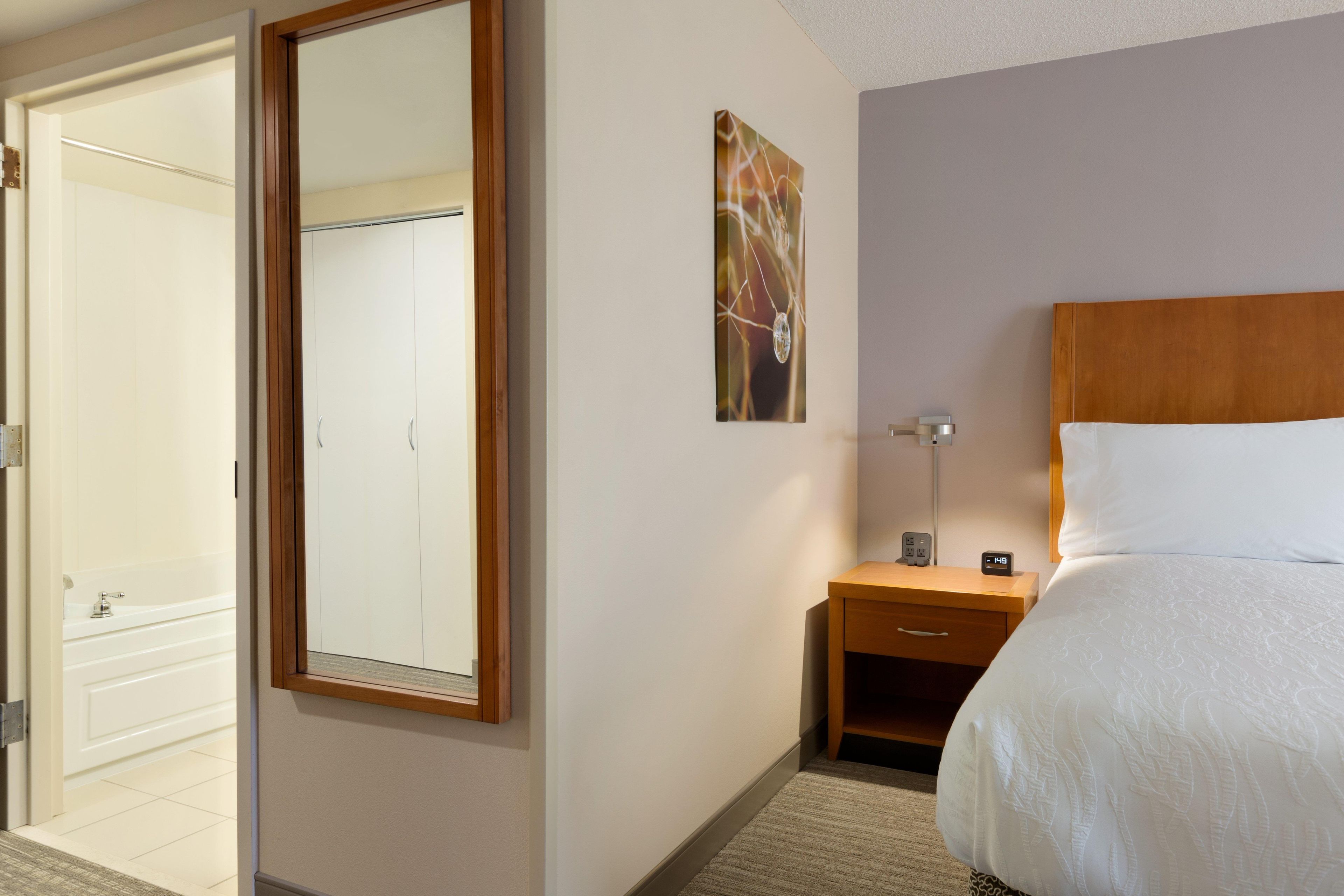 Habitación de hotel con cama, mesita de noche, espejo en la pared, un cuadro y baño con bañera.