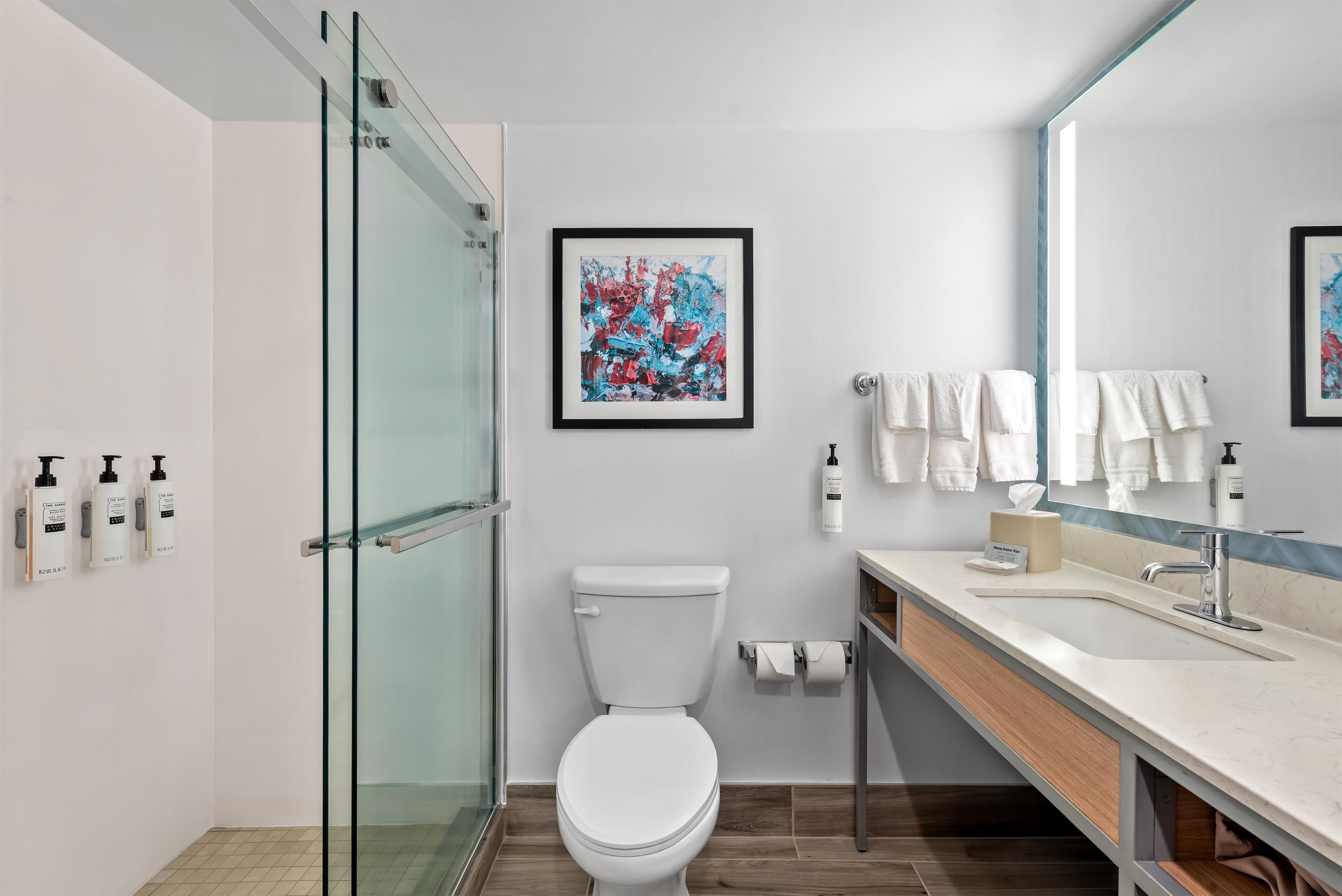A modern bathroom with a glass shower enclosure, a toilet, a sink with a mirror, and a framed abstract painting on the wall.