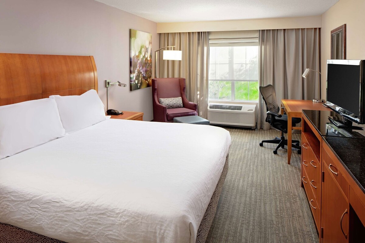 Una habitación de hotel con una cama grande, un escritorio con computadora, una silla, televisor y ventana con cortinas.