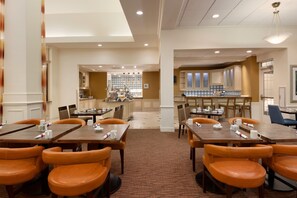 A hotel dining area with multiple tables and chairs, a kitchen in the background, and a bar area. - Hilton Garden Inn Fredericksburg (Fredericksburg)