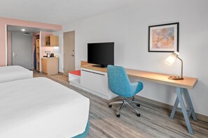 Habitación de hotel con dos camas, un escritorio con televisor, una silla, una lámpara de escritorio y un cuadro en la pared.