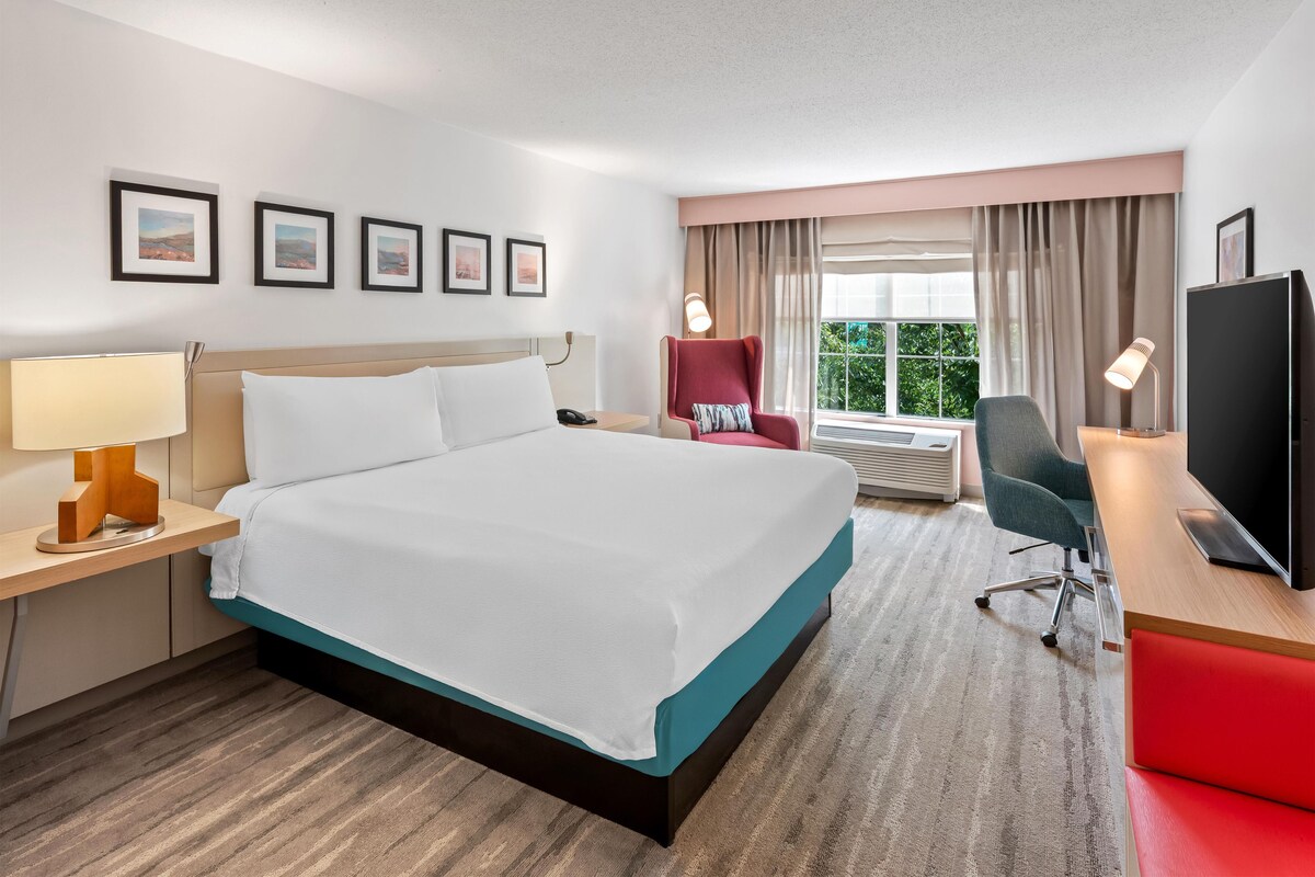 A hotel room with a bed, a desk with a computer, a chair, a TV, and a window with curtains.
