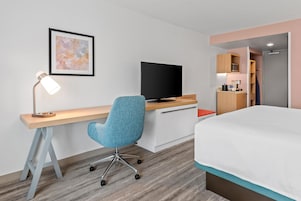 Une chambre d’hôtel comprenant un lit, un bureau avec une chaise, une télévision, un tableau encadré au mur et un coin cuisine.