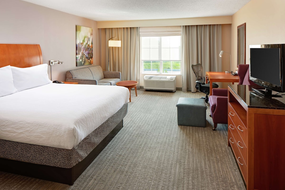 A hotel room with a large bed, a desk with a television, a chair, a sofa, and a window with curtains.