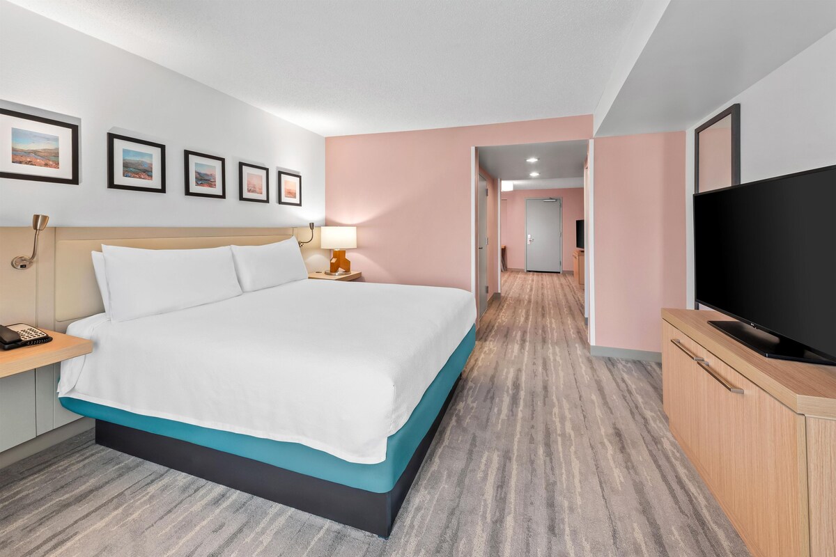 A hotel room with a bed, a TV, a desk with a lamp, and a hallway leading to another room.