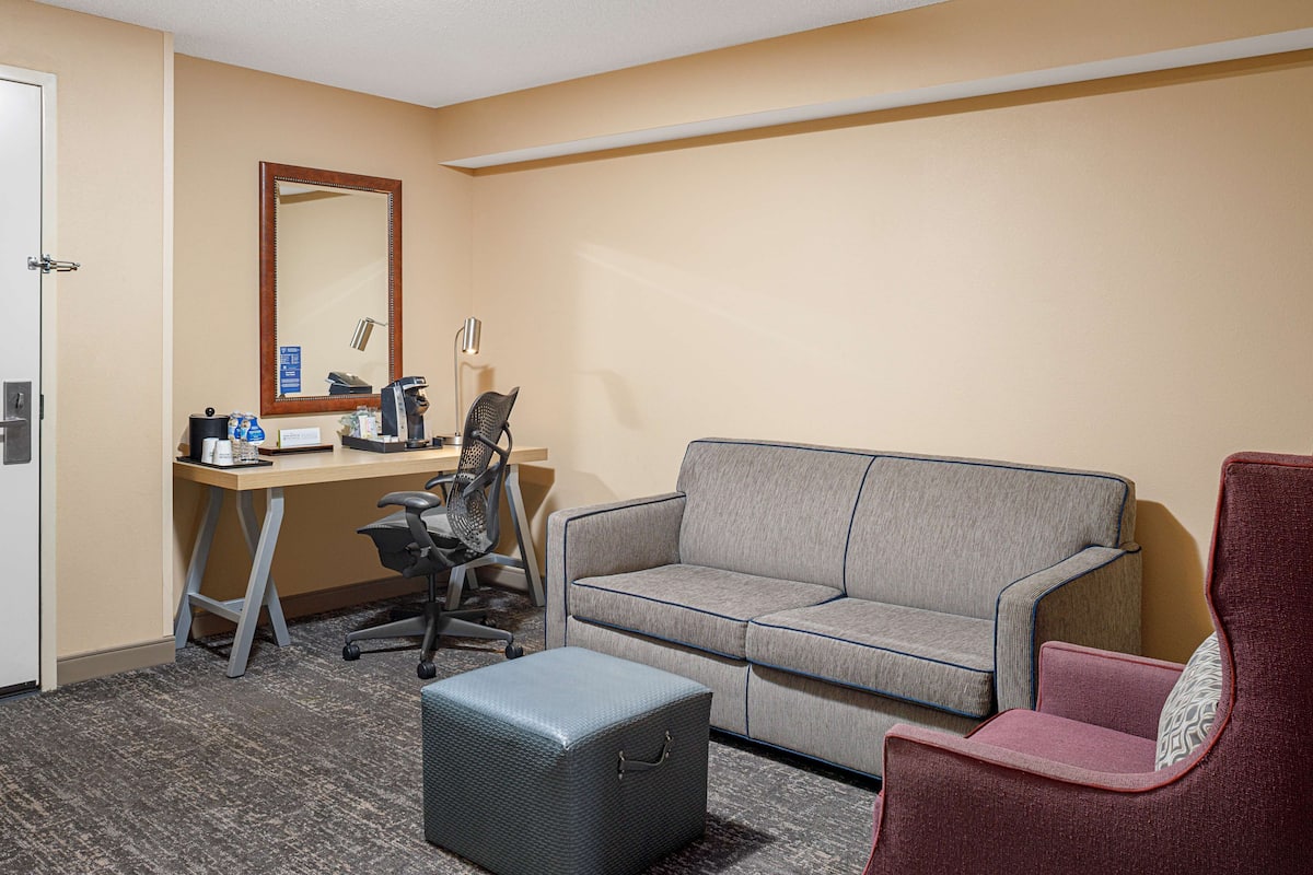A hotel room with a desk, a grey sofa, a red armchair, a small ottoman, and a mirror on the wall.