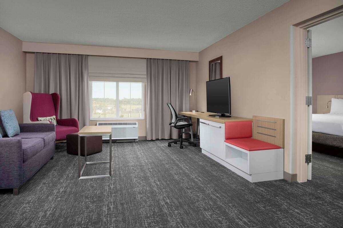 A hotel room with a sofa, a desk with a TV, a bed, and a view of the city.
