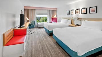A hotel room with two beds, a desk, a TV, and a view of greenery outside.